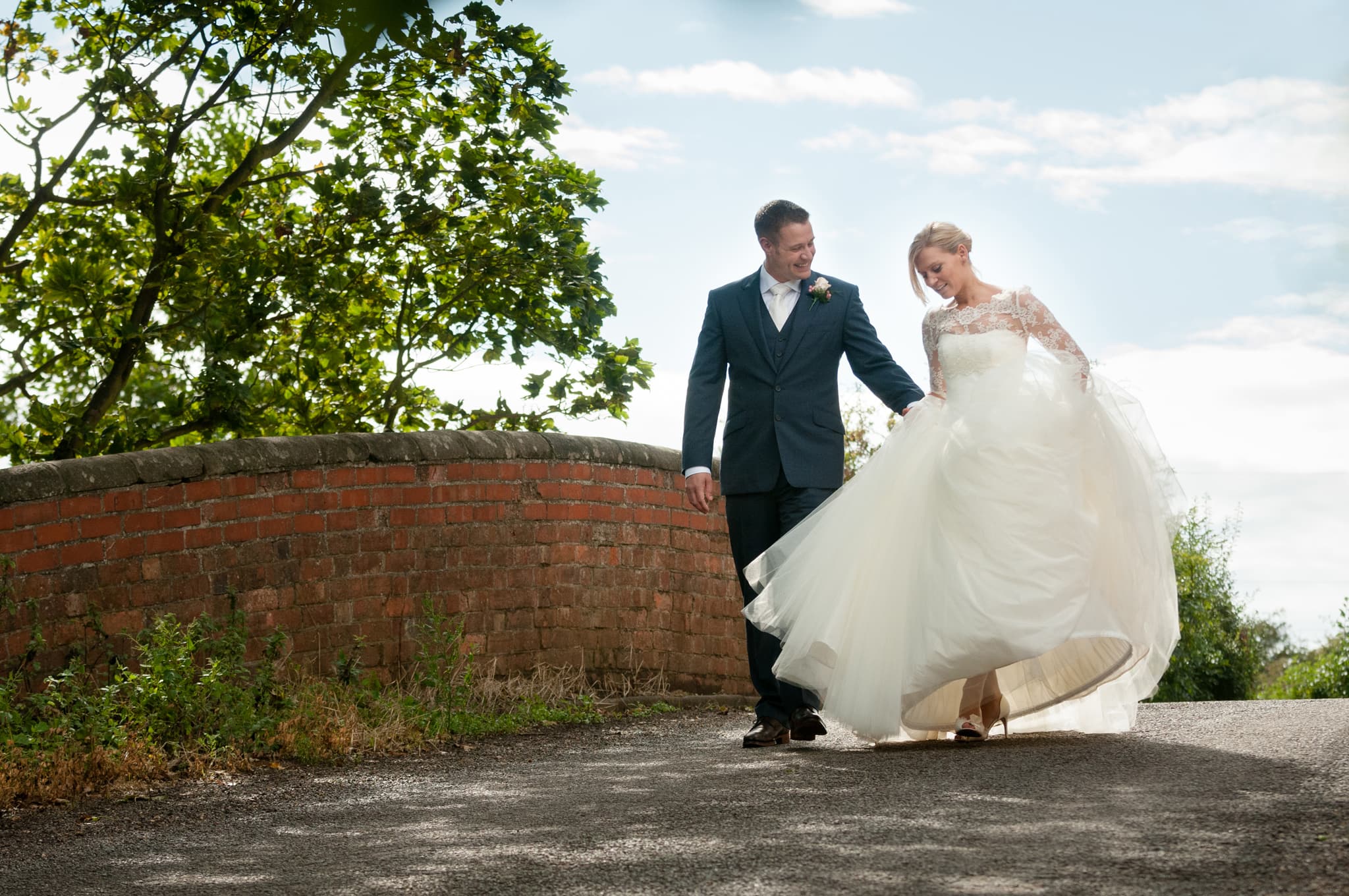 Groom helping the bride with her dress as they walk over a bridge with red brick walls outside Dodmoor House