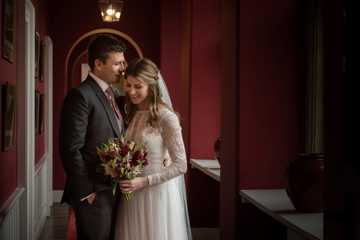 Portrait of a bride and groom in a deep red painted corridor which matches the bride's bouquet