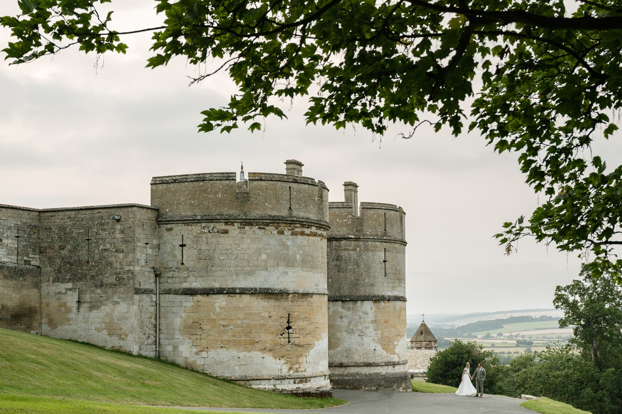 A bride and groom walking in front of the two towers at the entrance to Rockingham Castle