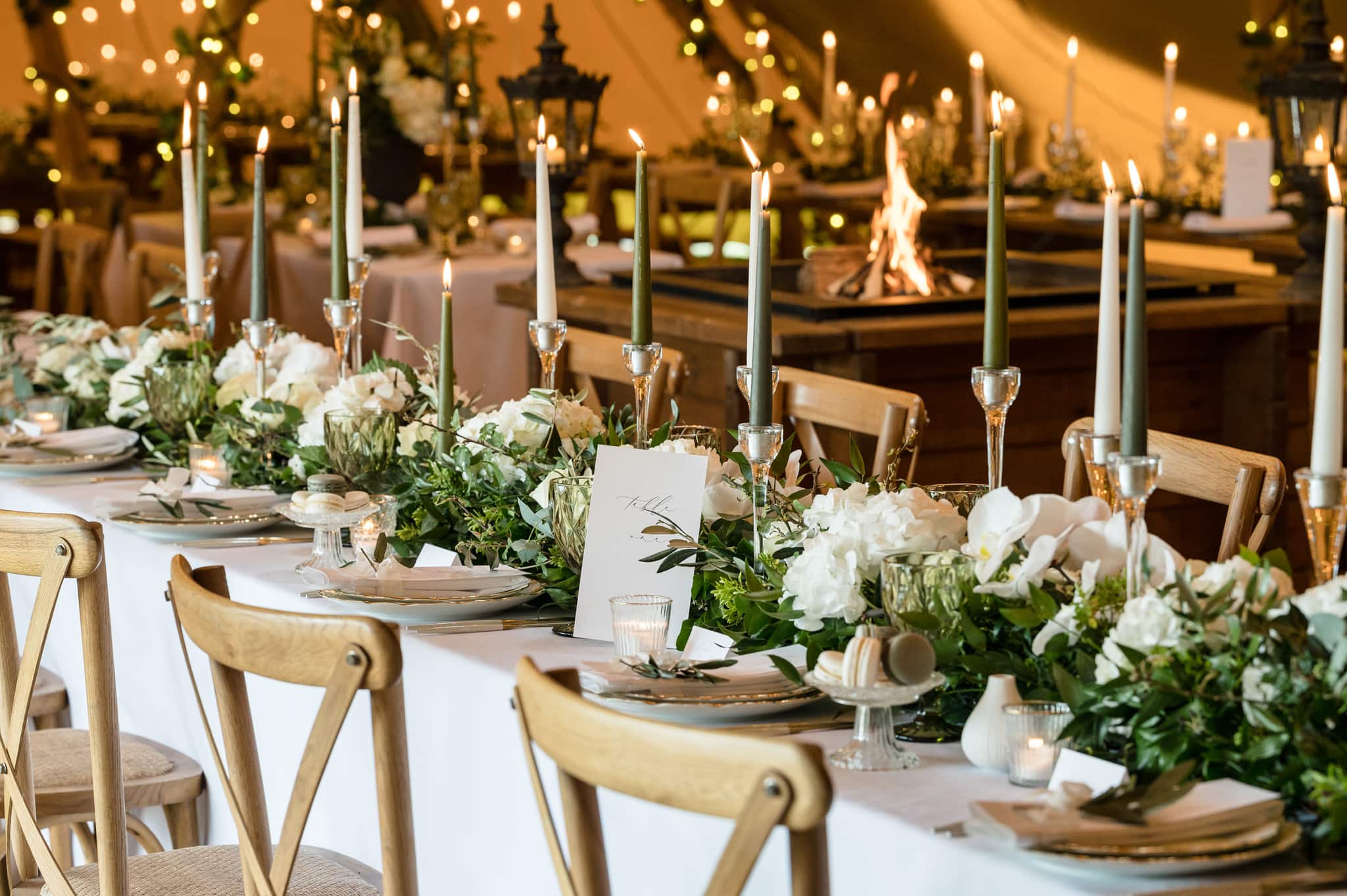 Long bench style table with oak cross-back chairs and white floral table decorations with candles in glass holders and foliage