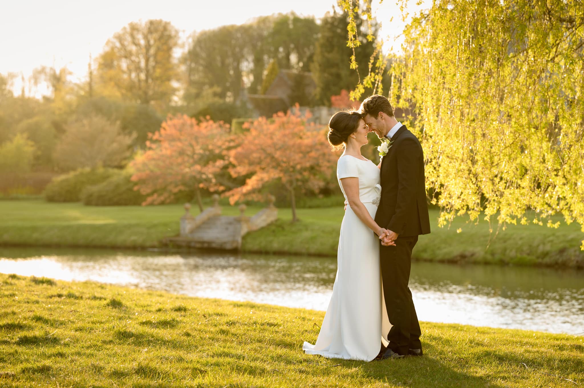 Bride and groom standing together by a willow tree on the riverbank at Deene Park during golden hour