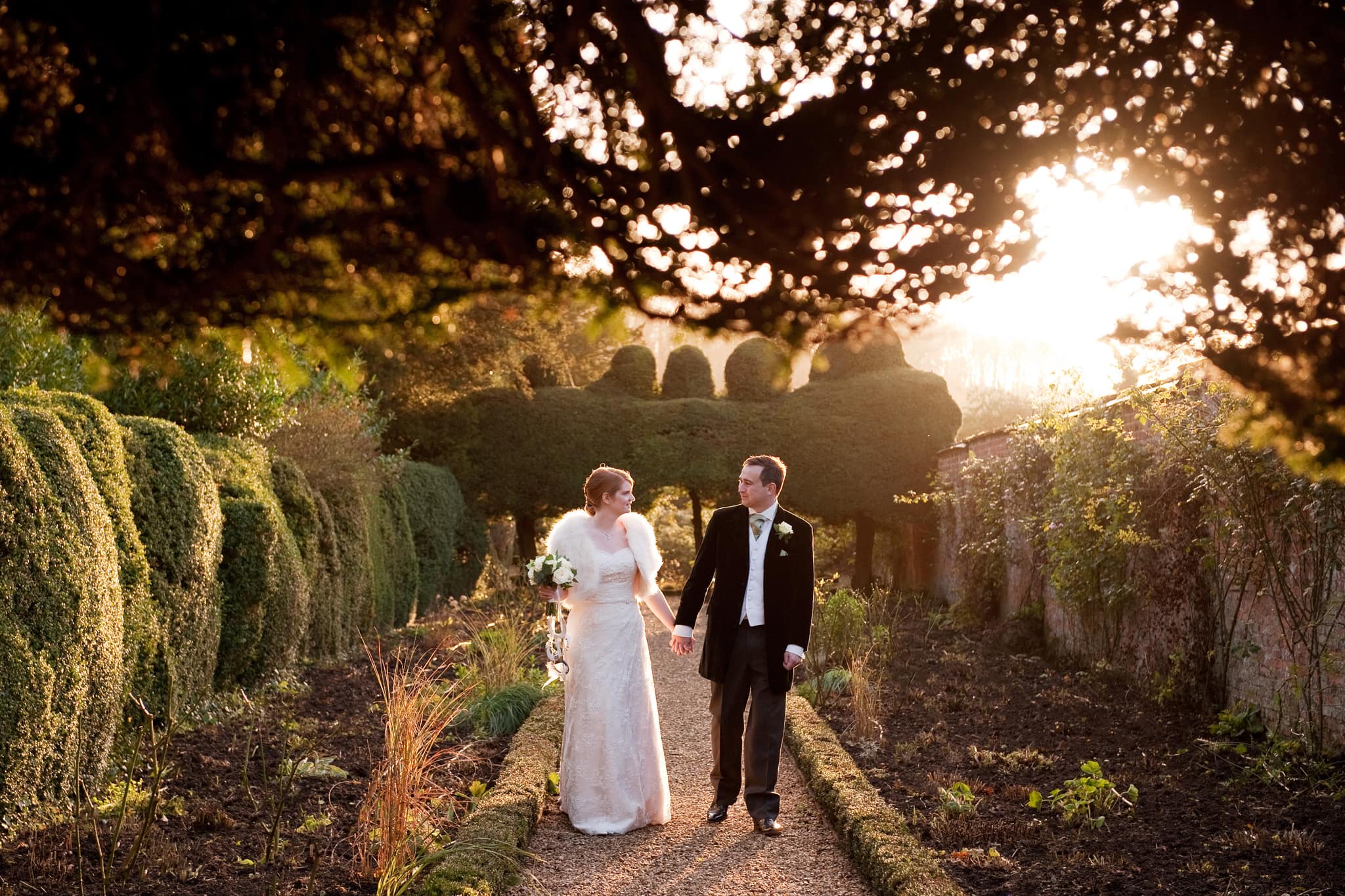 Couple walking down the long border in Kelmarsh Hall gardens lit by the golden hour sun behind them