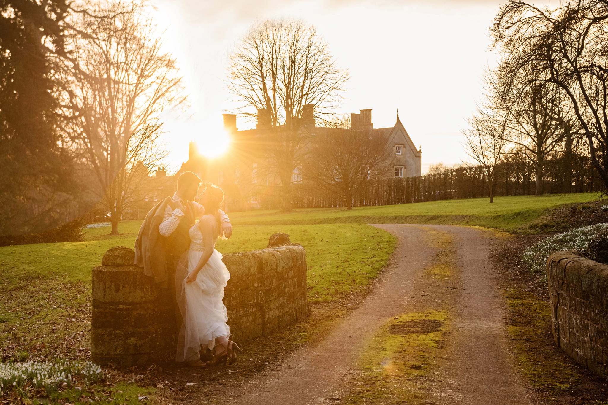 Bride and groom cuddling by a stone bridge with Weston Hall in the background and everything bathed in golden hour sunlight