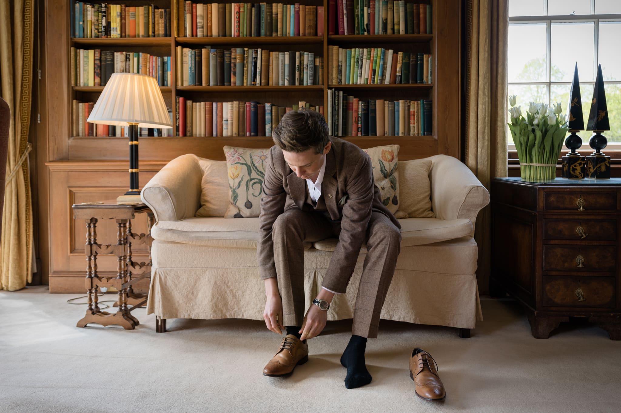 Groom sitting on sofa to put on his shoes in the library at Sutton Bonington Hall