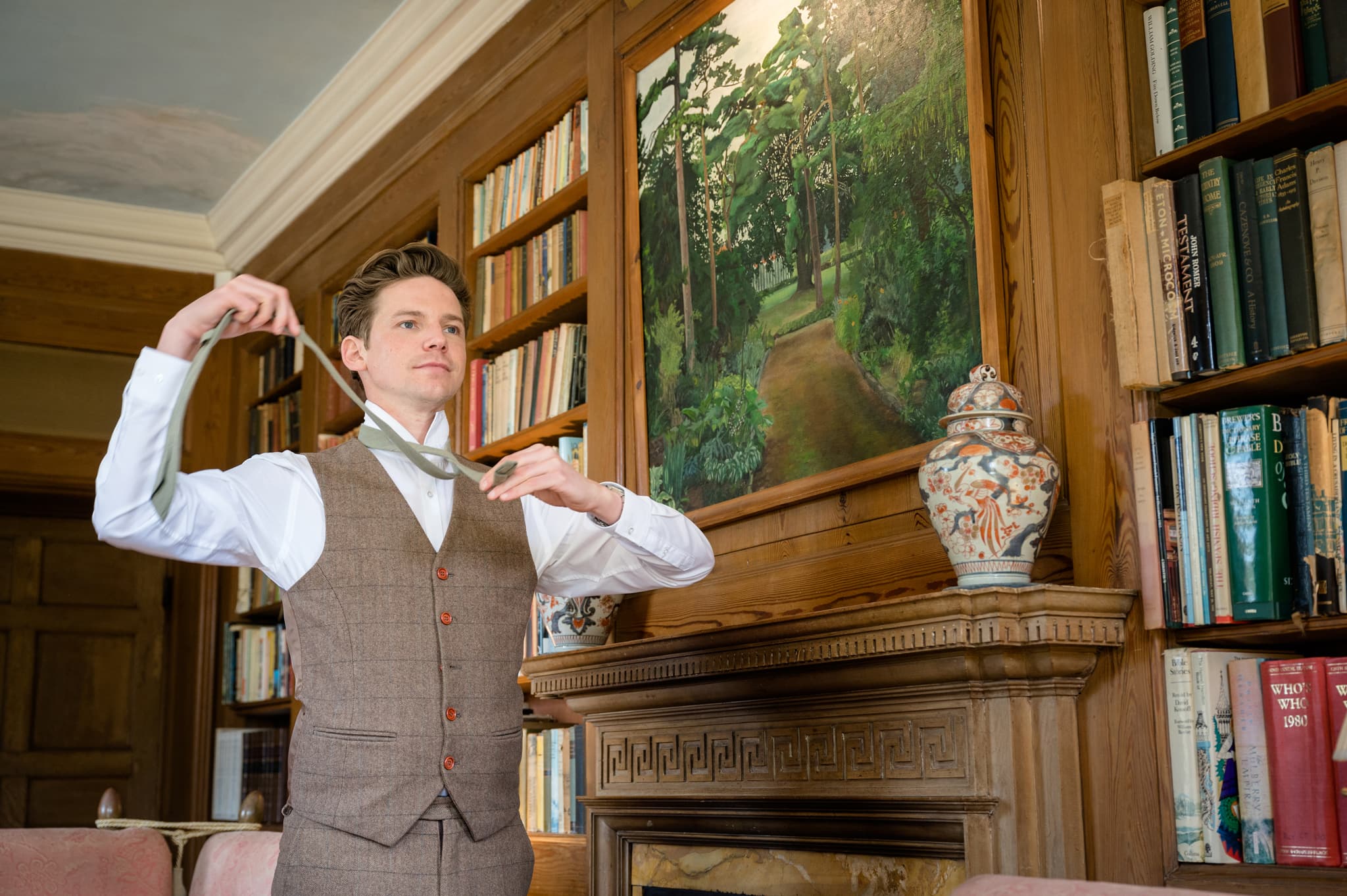 Groom putting on his tie in front of the fireplace in the library at Sutton Bonington Hall