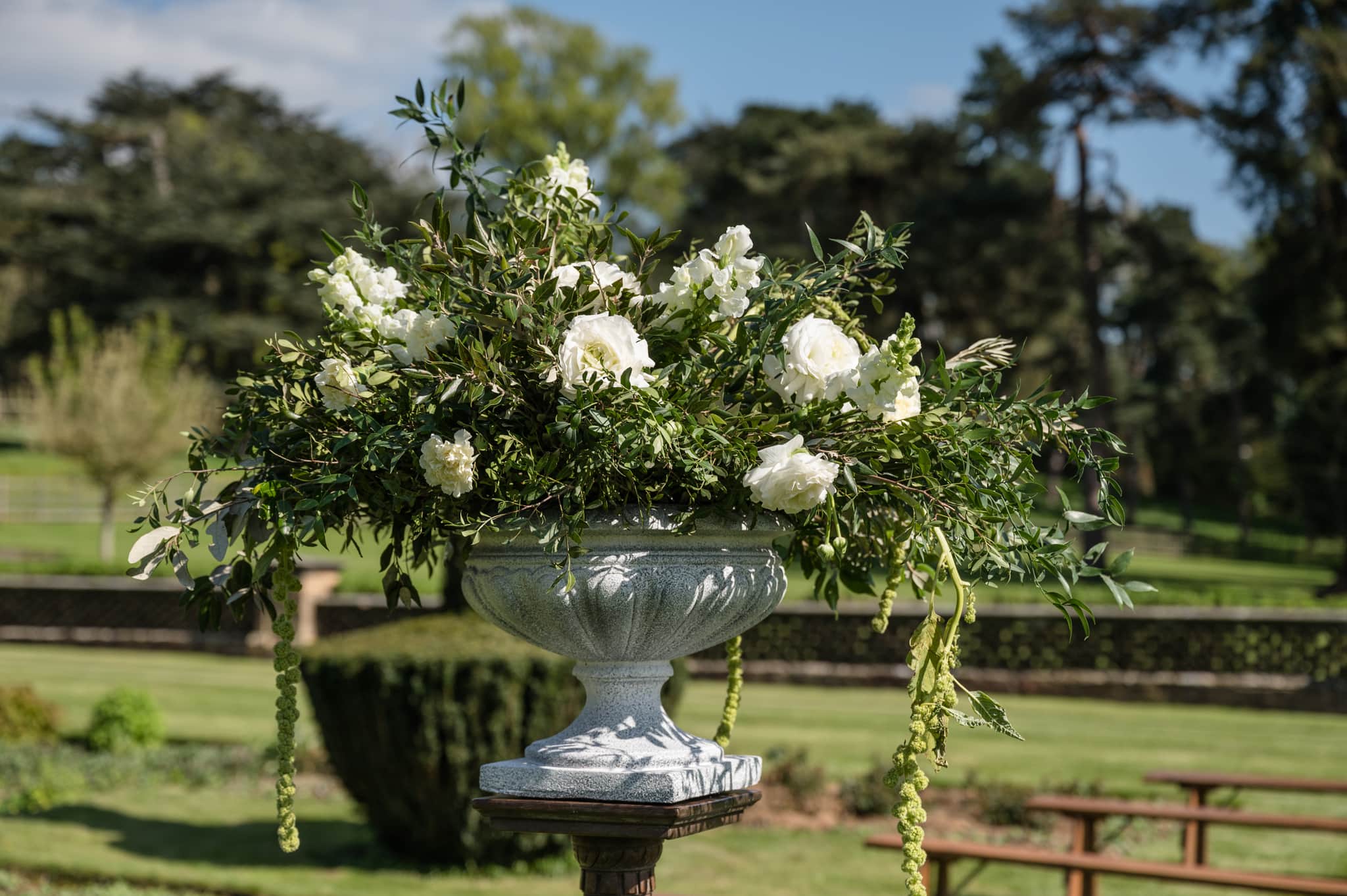 Close up of white flowers and greenery in an urn at the start of the aisle for an outdoor ceremony