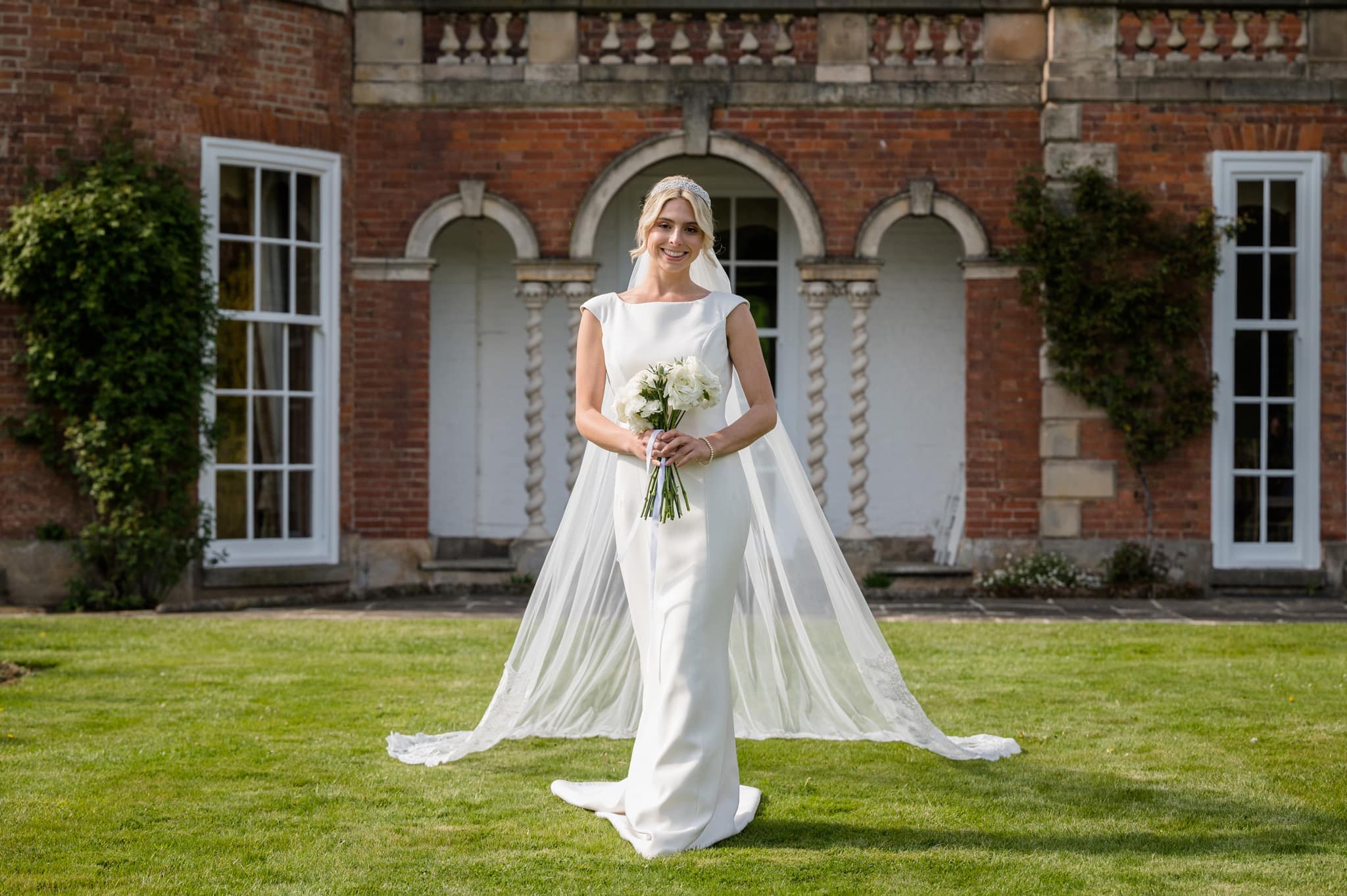 Bride in a simple white dress and long veil framed within an archway of Sutton Bonington Hall behind her