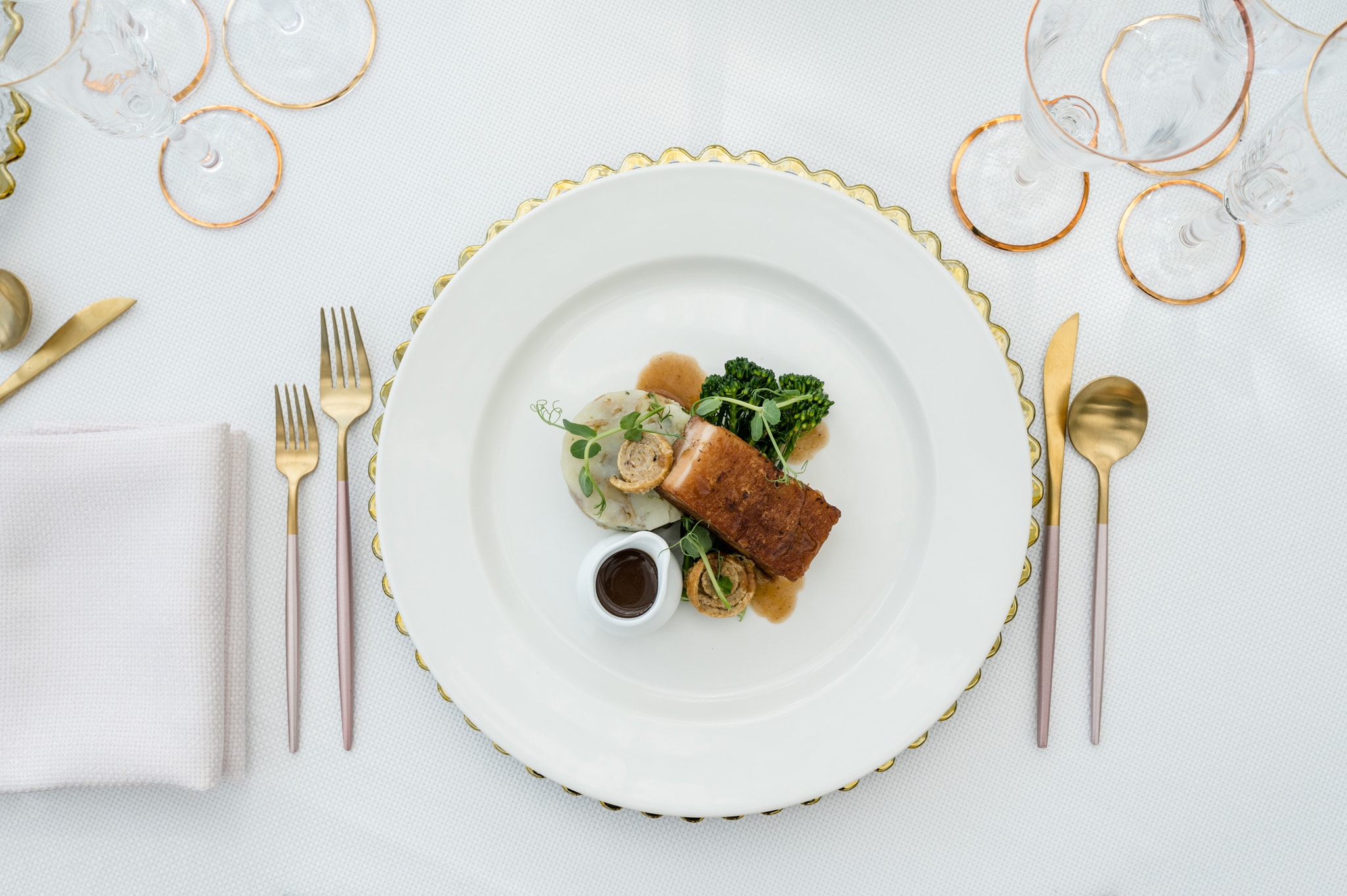 Pork belly wedding breakfast main course served on a white plate and gold scallop edge charger with frosted pink cutlery