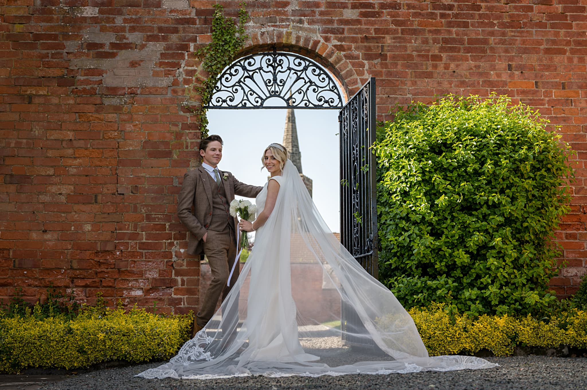 Bride and groom in front of a wrought iron gate with the spire of Sutton Bonington church in the background