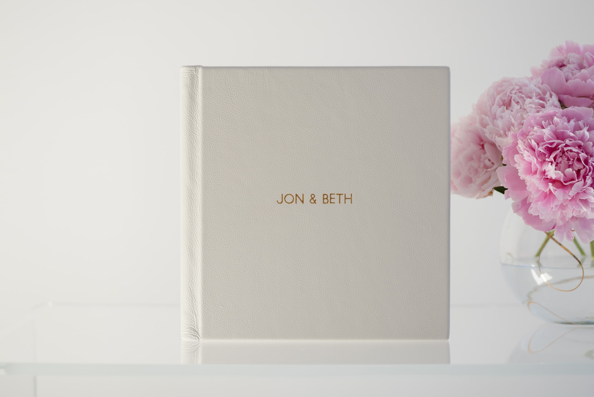 Pale grey square fine art wedding photo album propped up on a coffee table next to a vase of pink peonies