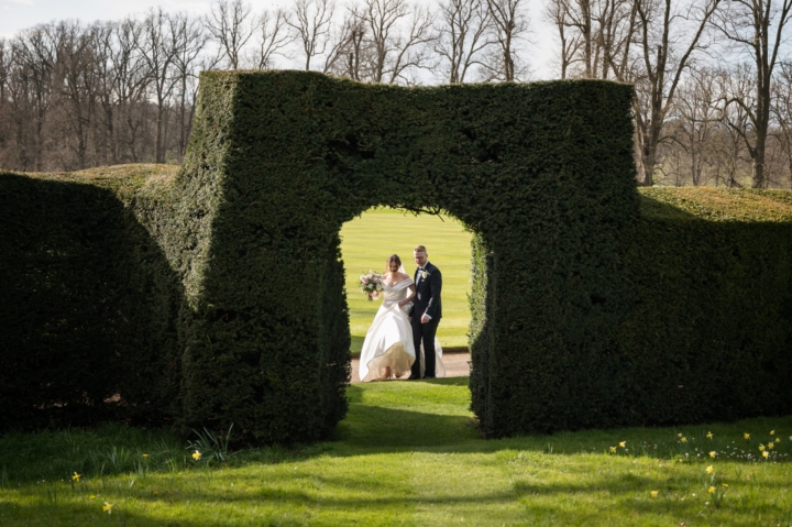 Bride and groom walking through gardens framed by a gap in the hedge at Boughton House