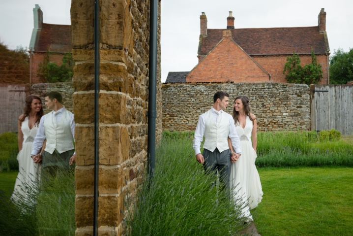 Bride and groom walking alongside a path of lavender with their reflection in a window at Dodford Manor