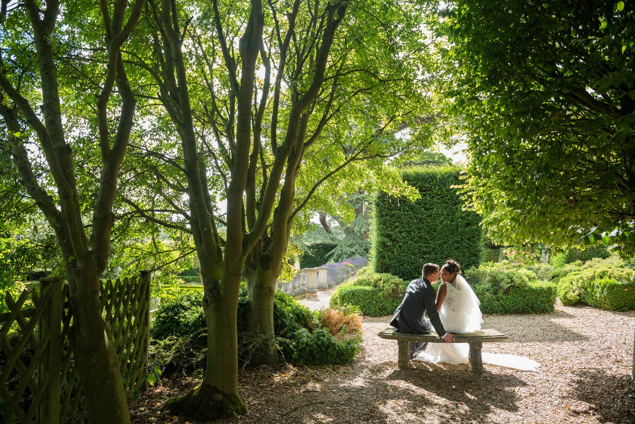 Bride and groom sitting on a bench under some trees at Fawsley Hall