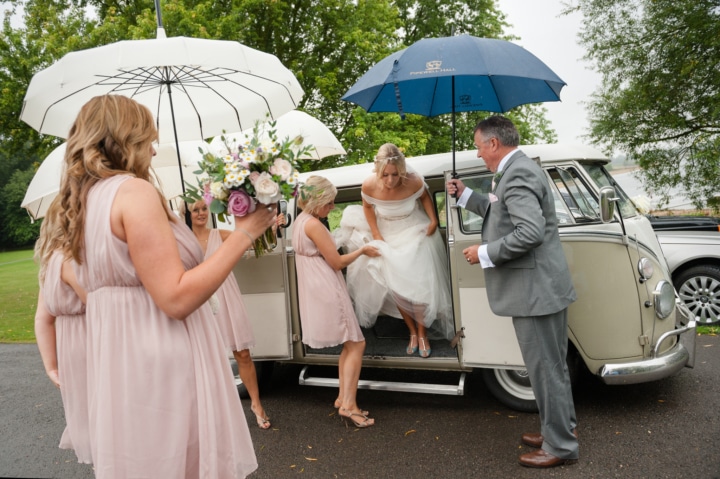 Bridesmaids protecting bride under umbrellas as she gets out of a campervan at Normanton church