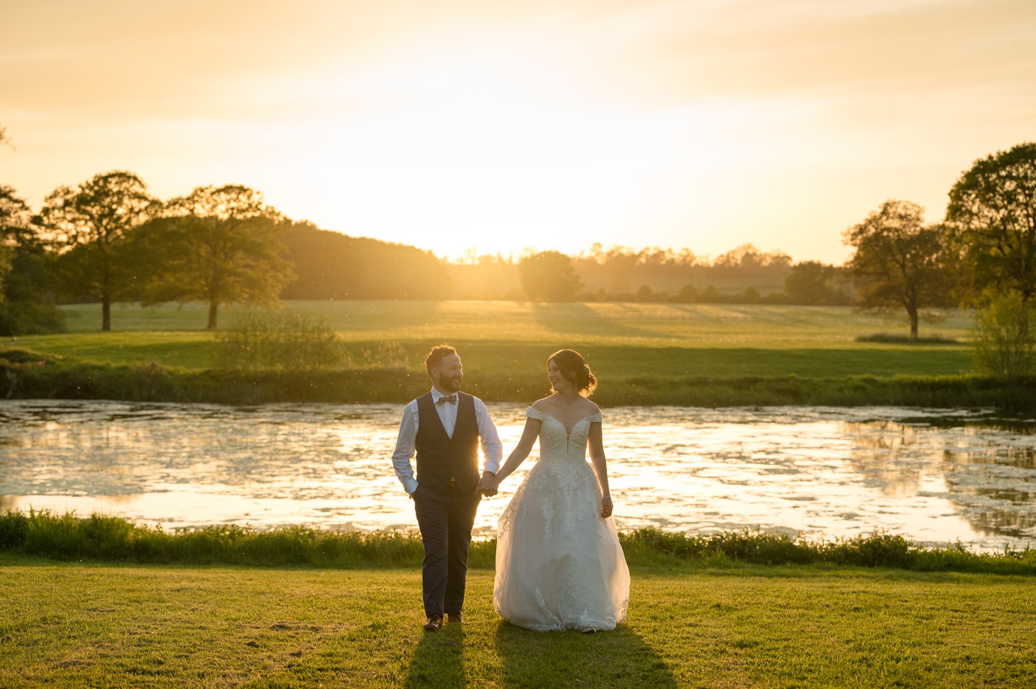 Bride and groom walking hand in hand through a field during golden hour with a countryside view and lake behind them