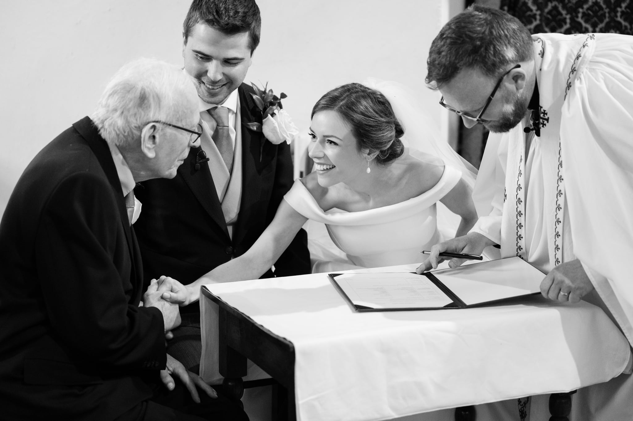 Bride leaning over to hold her Grandad's hand as the vicar signs the register