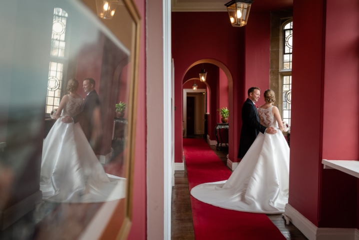 Bride and groom standing in a bay window with a reflection of them in a glass frame on the wall at Rushton Hall