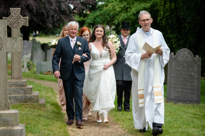 Vicar leading the bride and her dad down the path at Rushton church