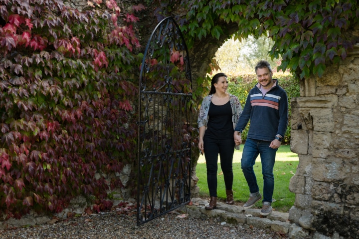 Engaged couple walking through an arched wrought iron gate at Notley Abbey