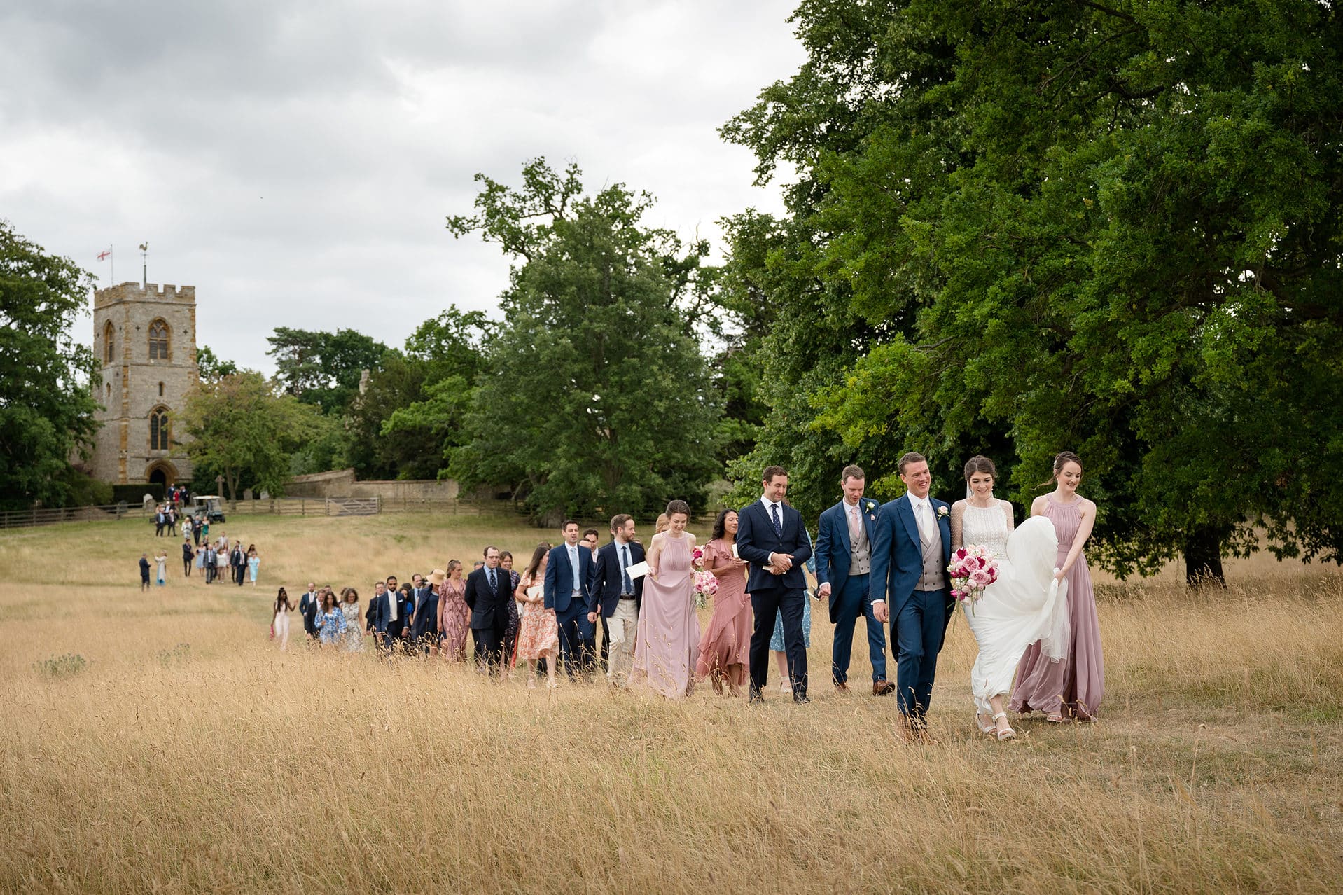 Bride and groom leading their wedding guests through a field of long grass with the church behind them