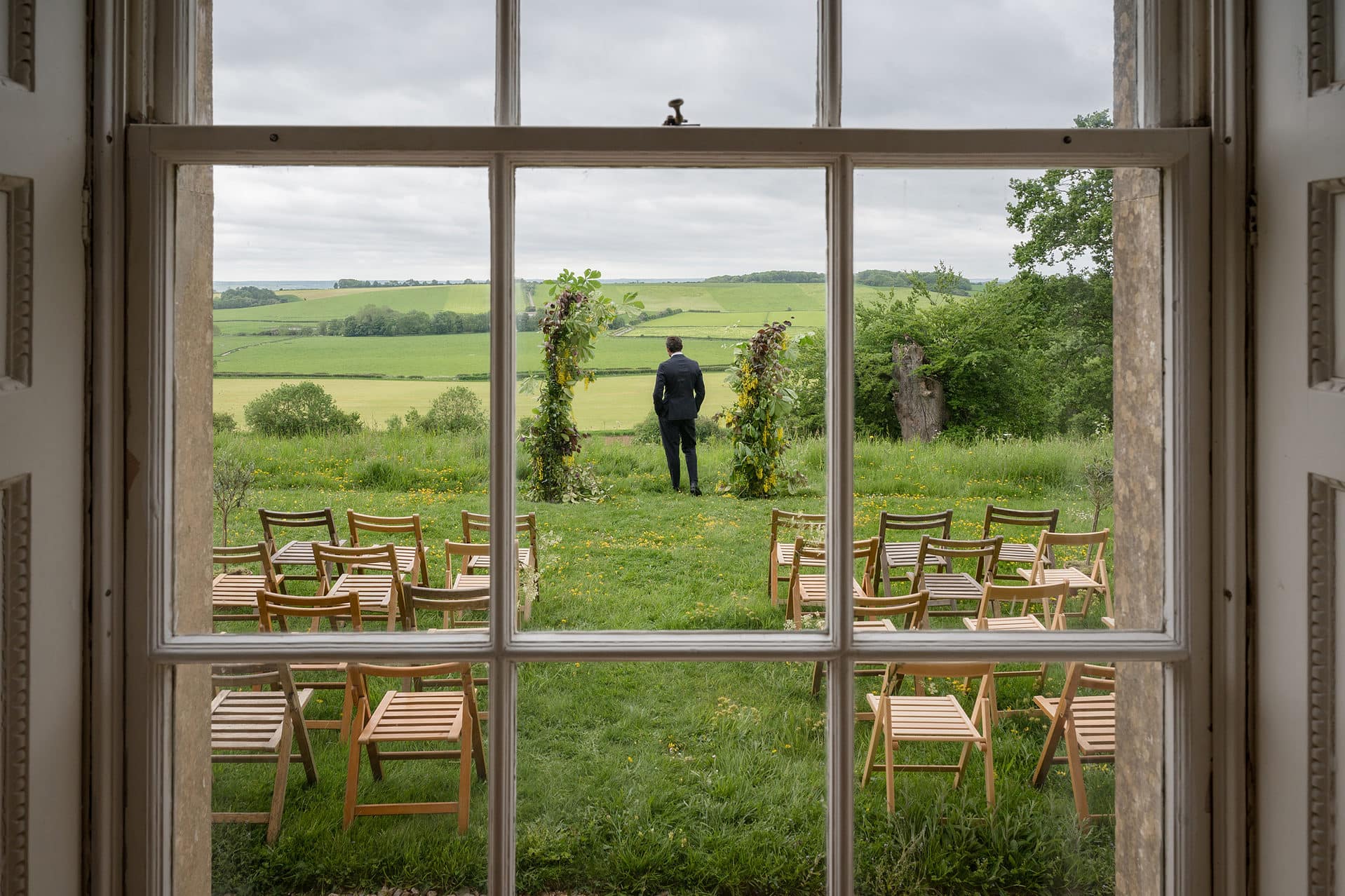 Shot through a window of a groom standing at the front of the outdoor ceremony area looking out over the Leicestershire countryside