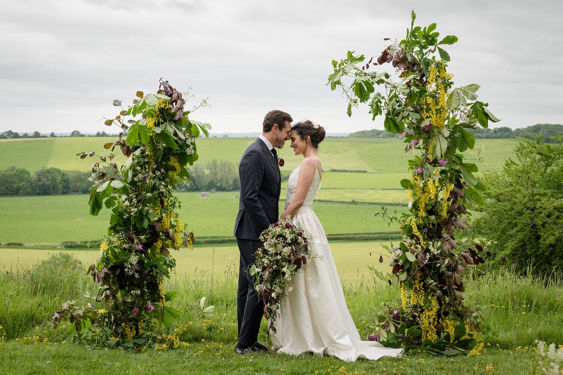 Bride and groom stood together between a deconstructed floral arch with views of rolling hills in the background