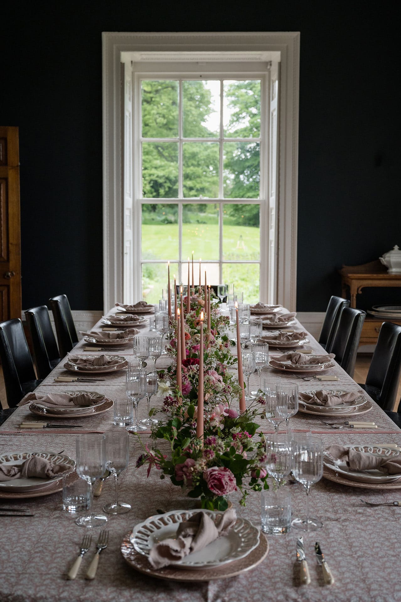 A long banqueting table set for dinner with pink flowers and tableware in a dining room with navy painted walls