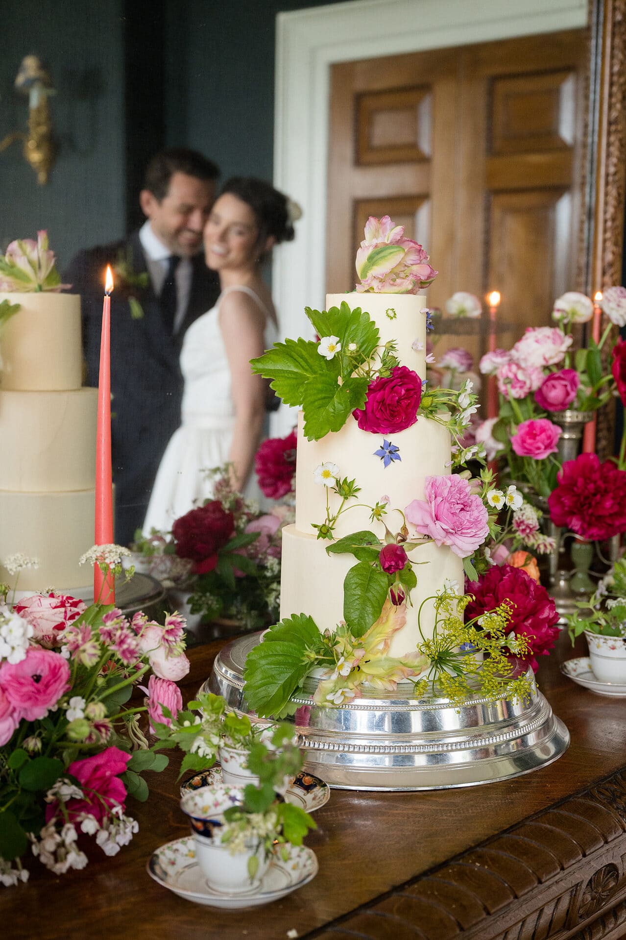 A buttercream wedding cake decorated with fresh flowers in front of a mirror which is reflecting the bride and groom