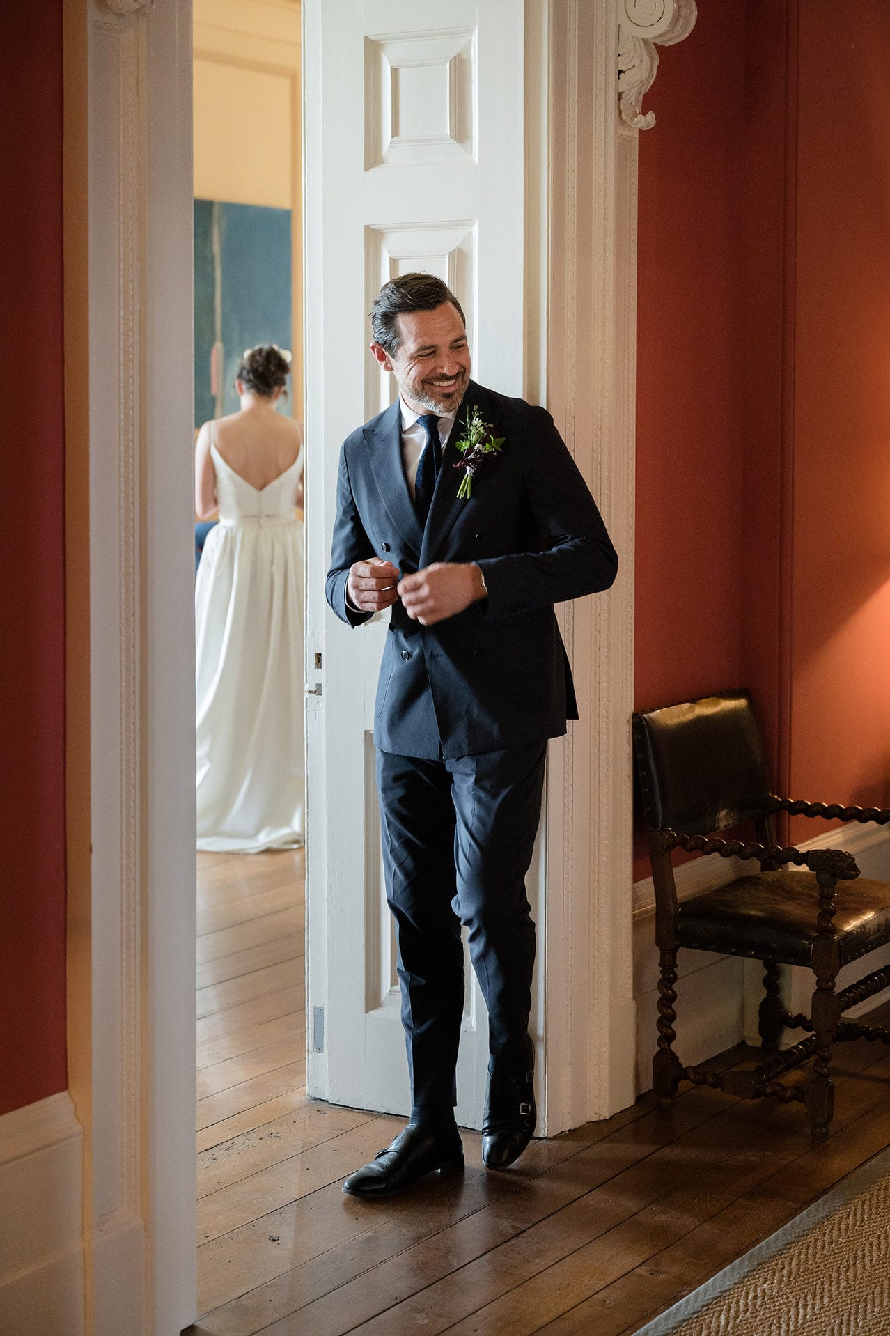 Groom standing in a doorway doing up his jacket with the back of the bride in the background