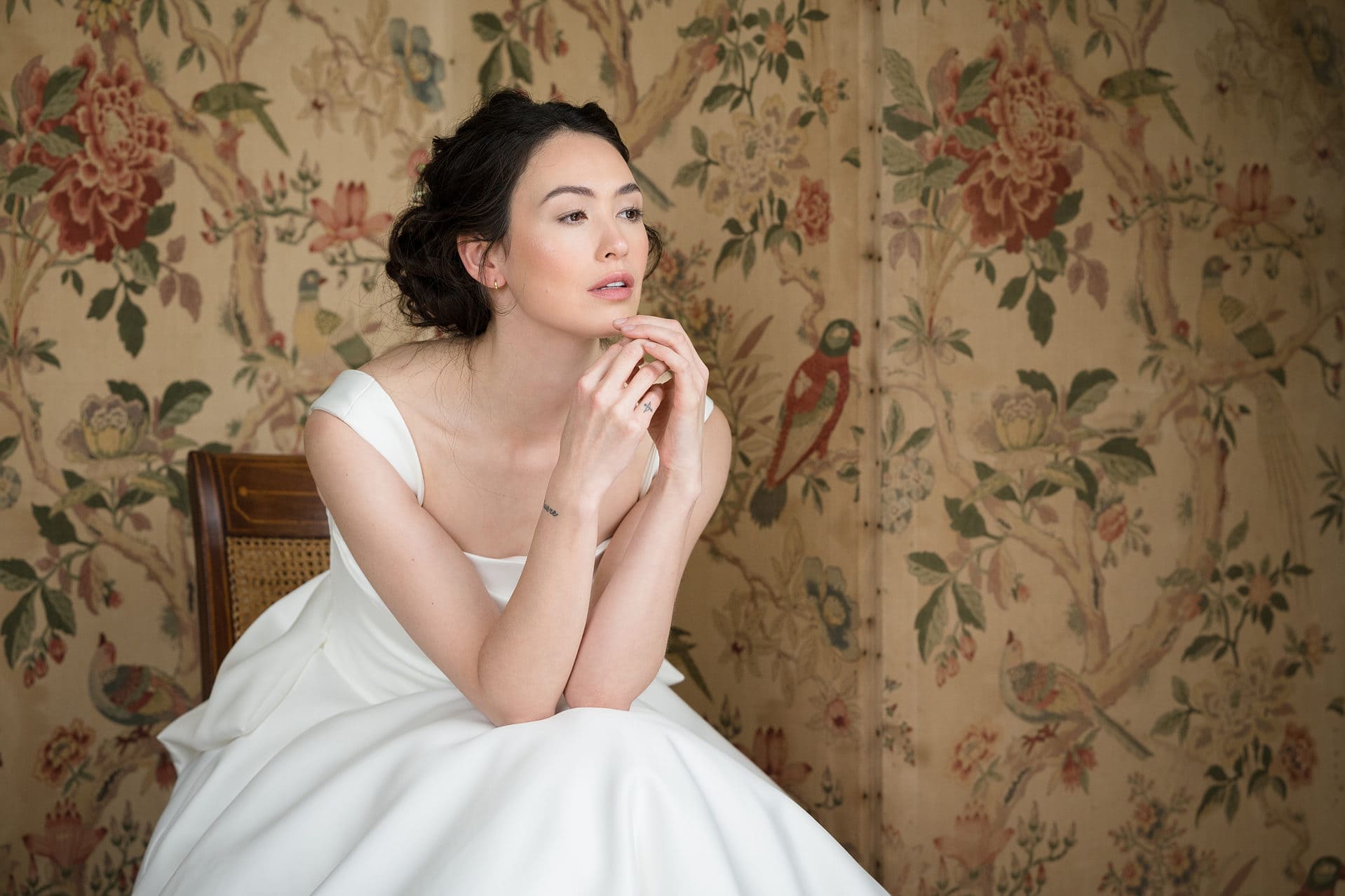 A bride sitting on an antique chair in front of a folding screen which is covered in a floral/fauna fabric
