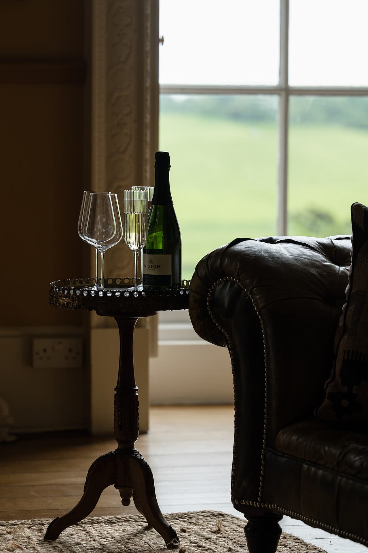 A bottle of wine and four glasses on an antique wine table next to the arm of a leather chesterfield sofa