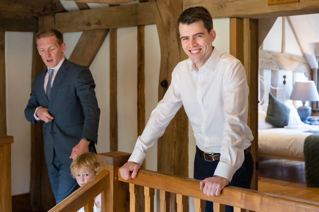 The groom leaning on a handrail and smiling at the camera whilst a flower girl is helped down the staircase by her dad