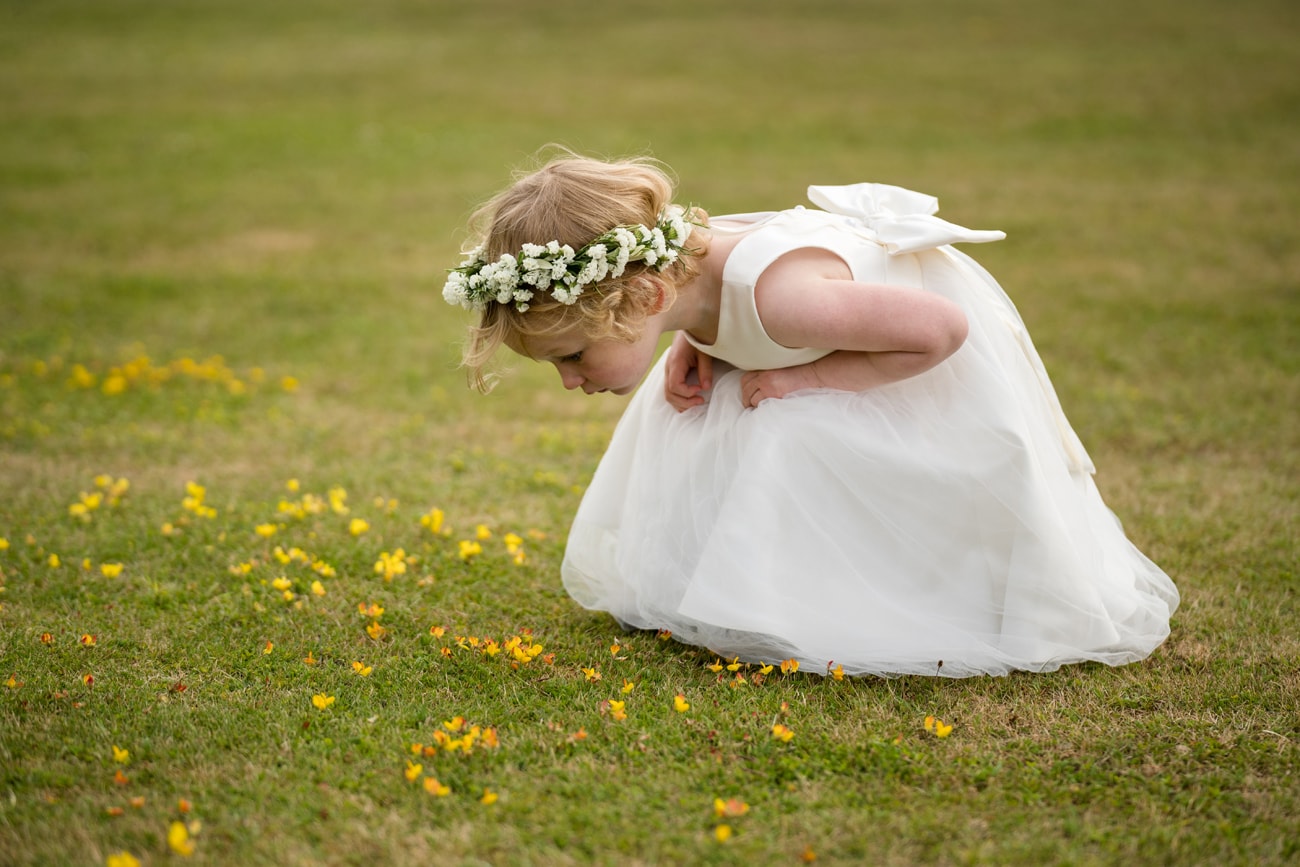 A flower girl in a white dress and flower crown bending down to look closely at buttercups in the lawn