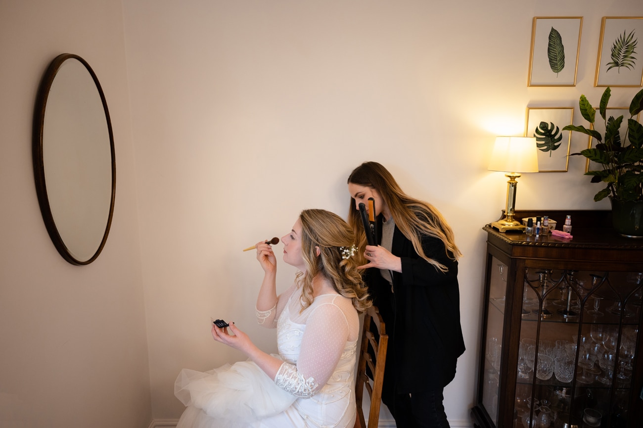 Bride finishing her makeup in a mirror while the hair stylist finishes her hair
