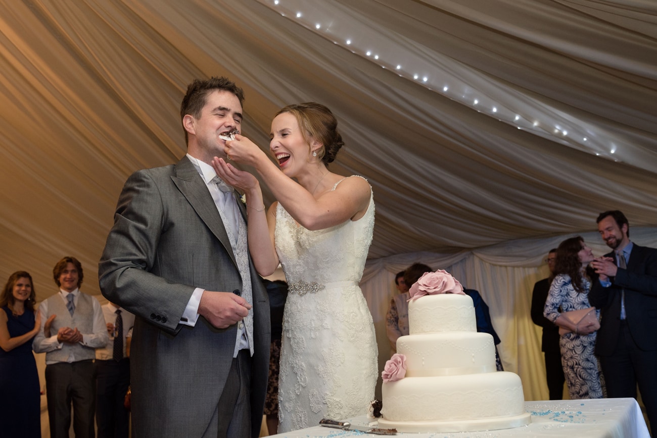 Bride laughing as she feeds the groom with a piece of wedding cake
