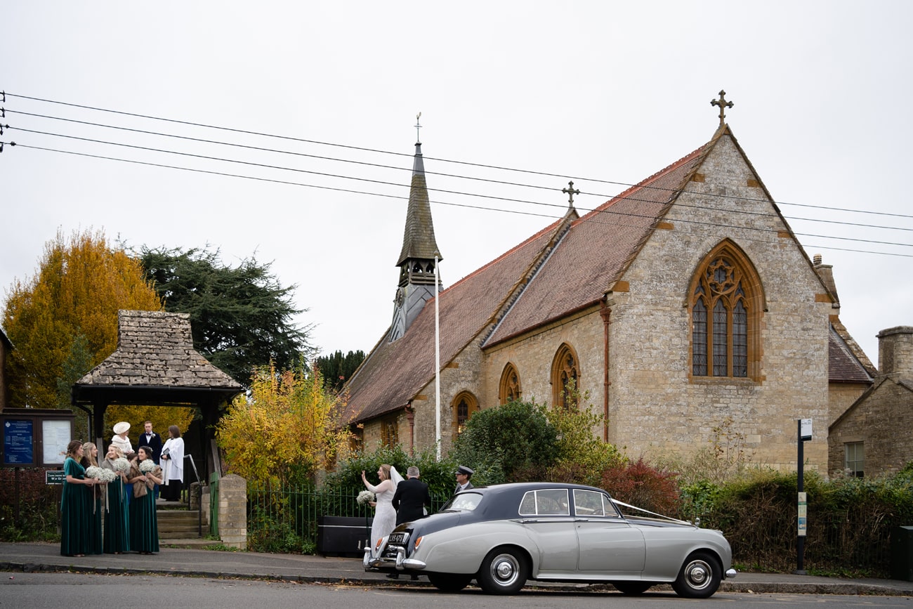 Bride next to wedding car waving to her bridesmaids as she arrives at St Michael's church in Silverstone