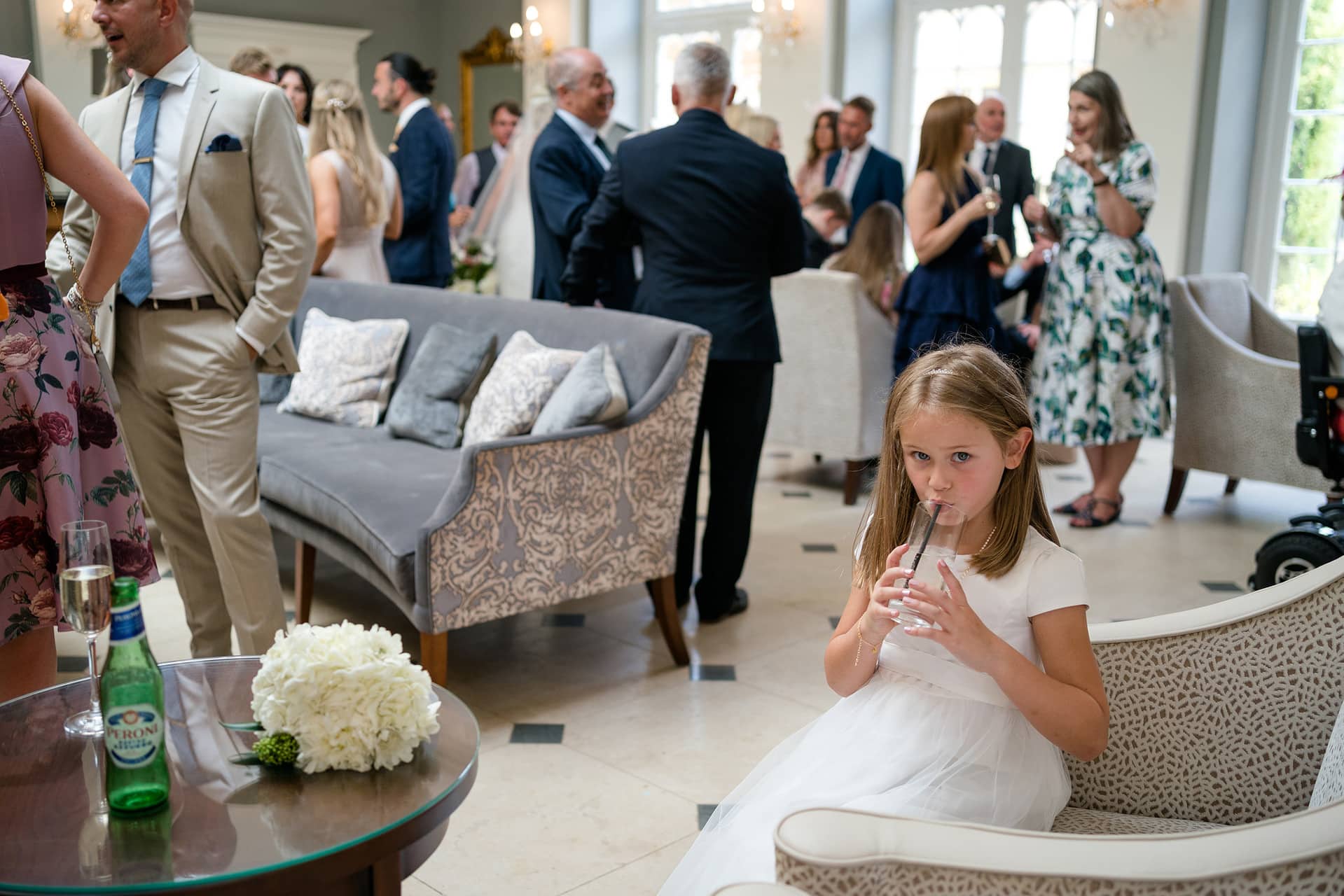 Flower girl drinking through a straw amidst a busy drinks reception behind her
