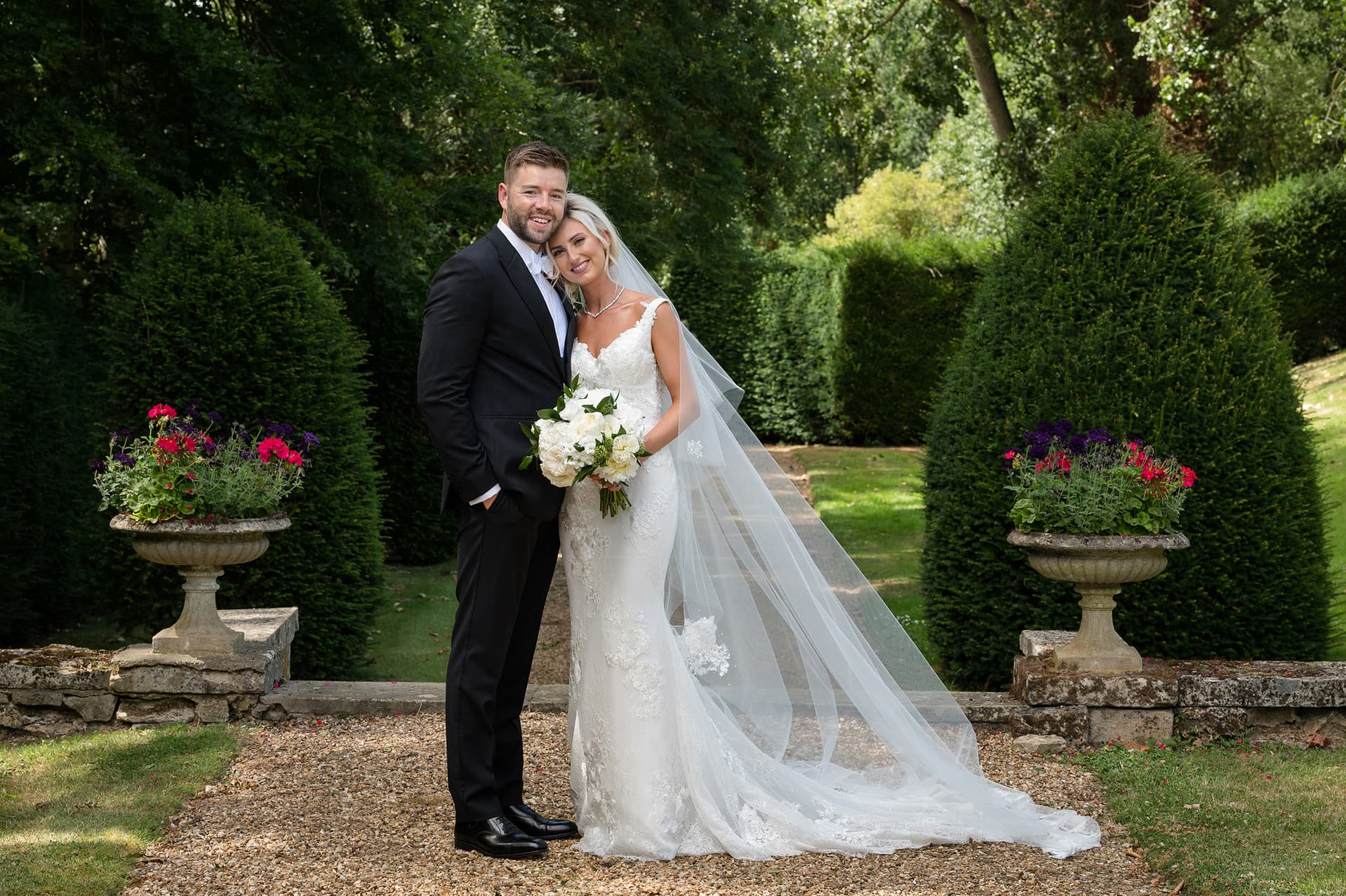 Posed portrait of bride and groom looking at camera in front of a background of trees and hedges