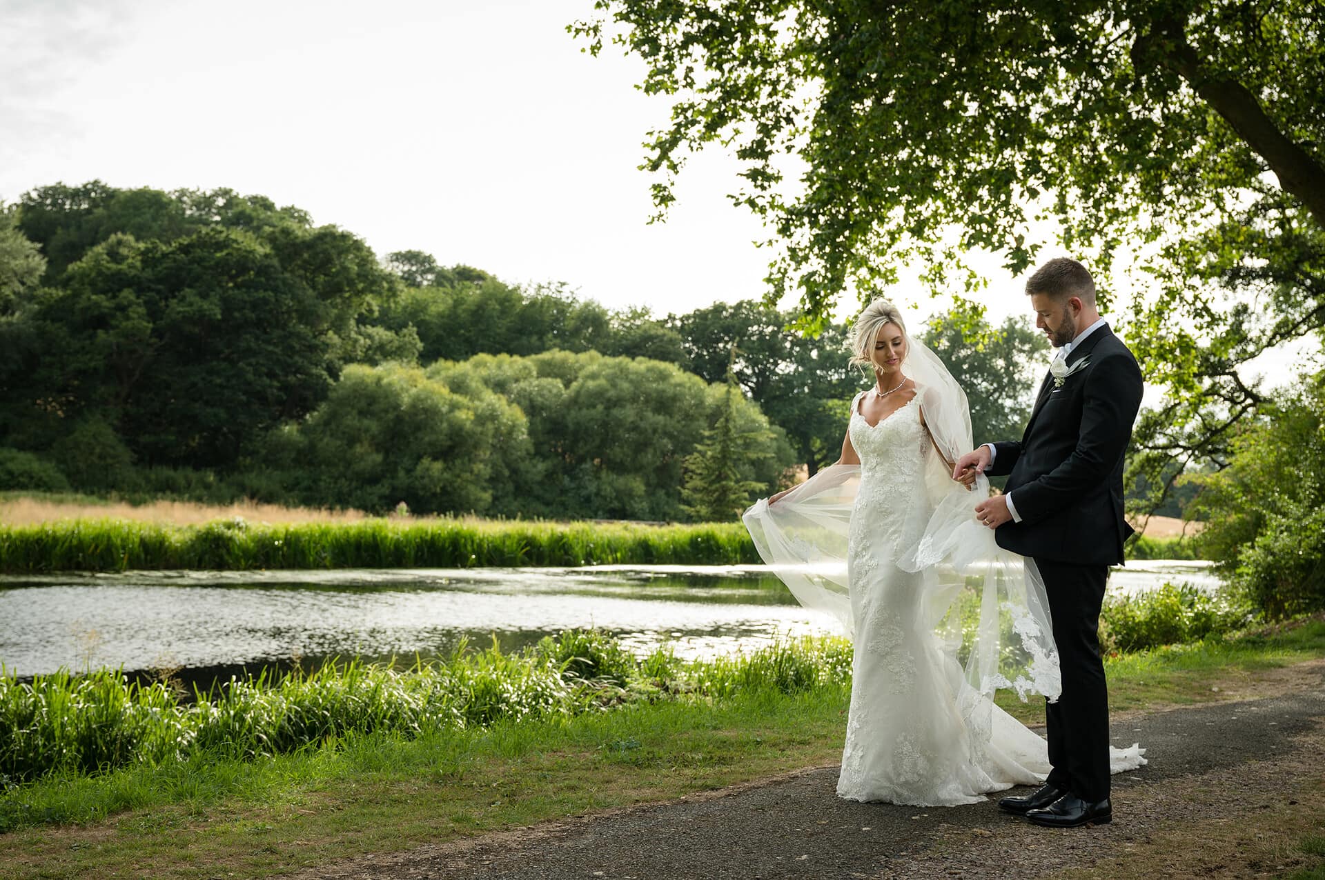 Groom helping bride with her veil as they walk along a lakeside path