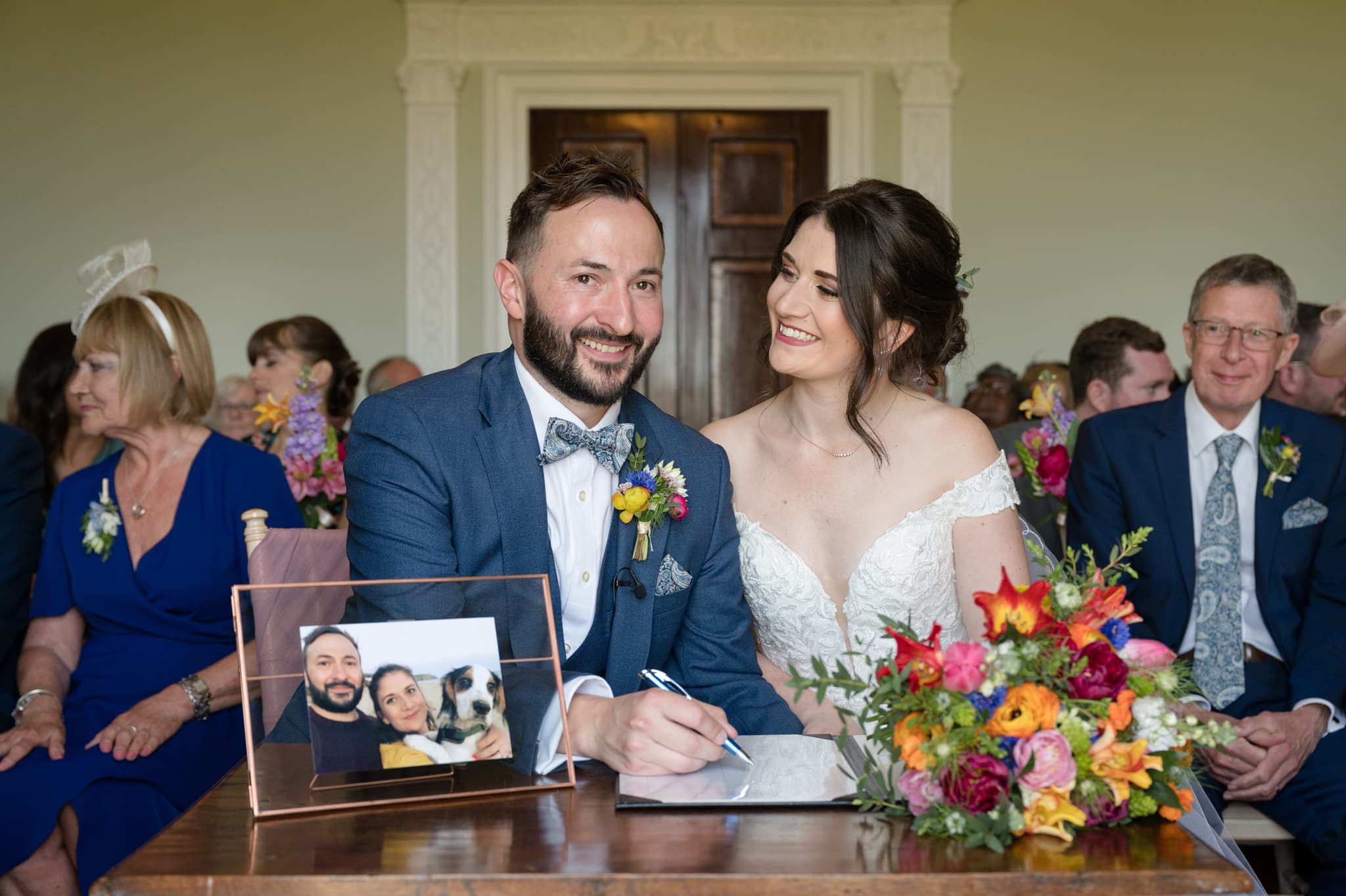 Bride and groom signing the register - with a photo of the couple and their dog on the table alongside the bride's bouquet