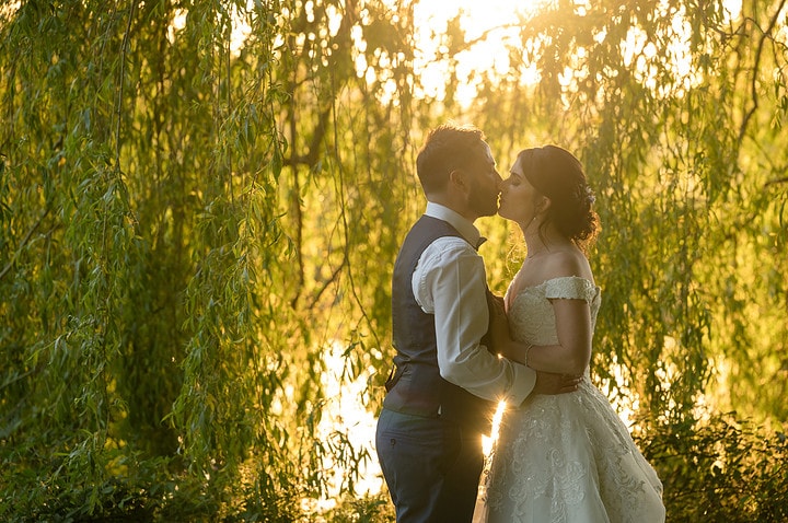 Bride and groom about to kiss under willow trees with golden hour sunlight shining through the leaves behind them