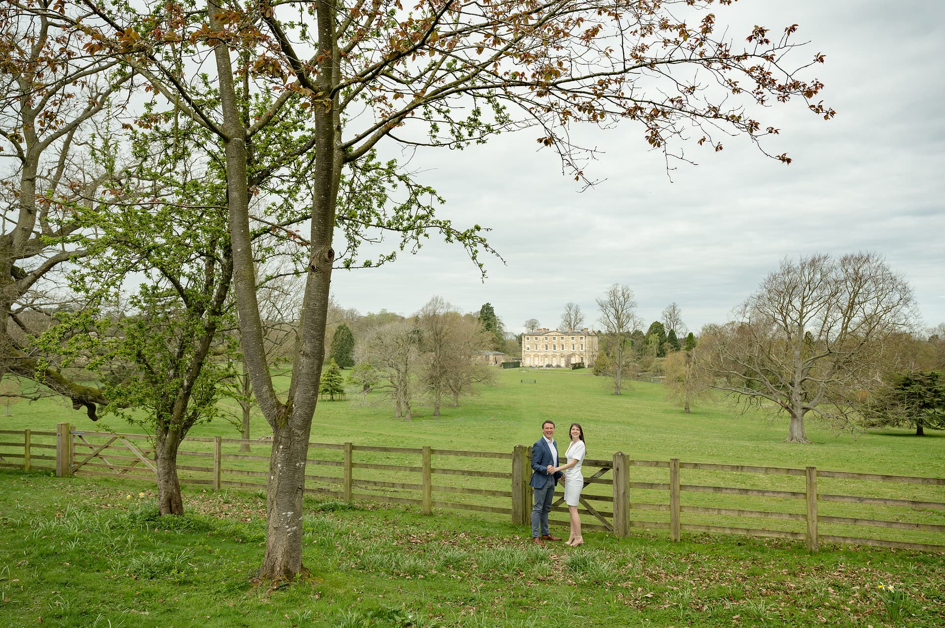 Engaged couple standing by the gate at Courteenhall church with Courteenhall house behind them