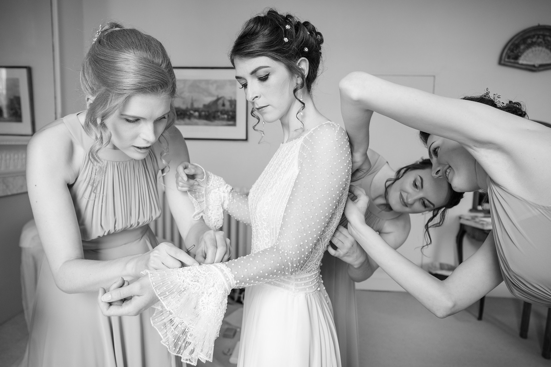 One bridesmaid doing up buttons on bride's sleeve while two other bridesmaids do up the buttons on the back of her dress