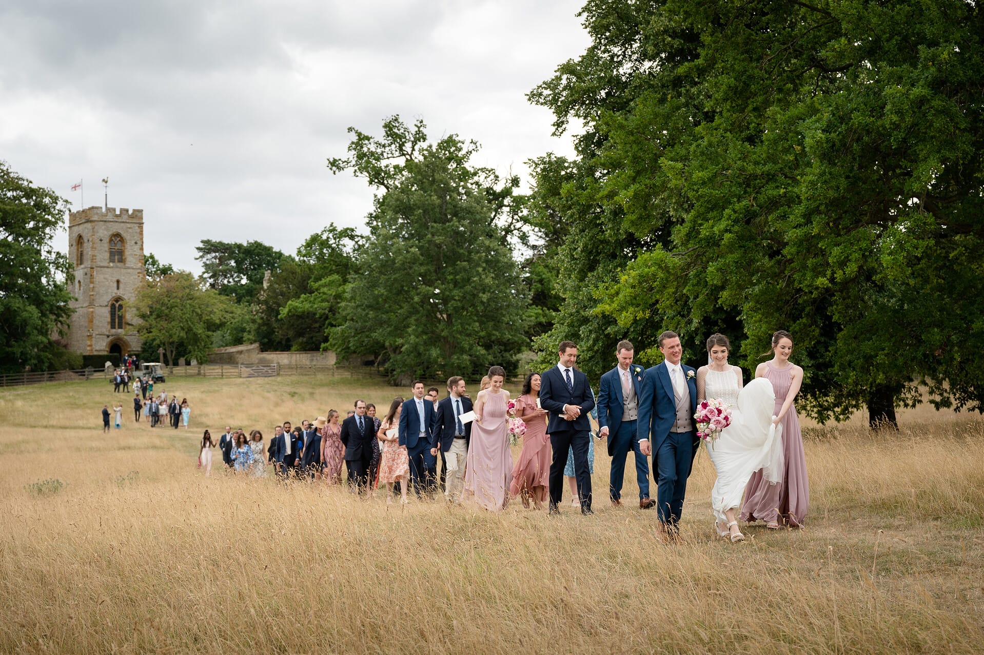 Bride and groom leading their wedding guests from the church through a field of long grass