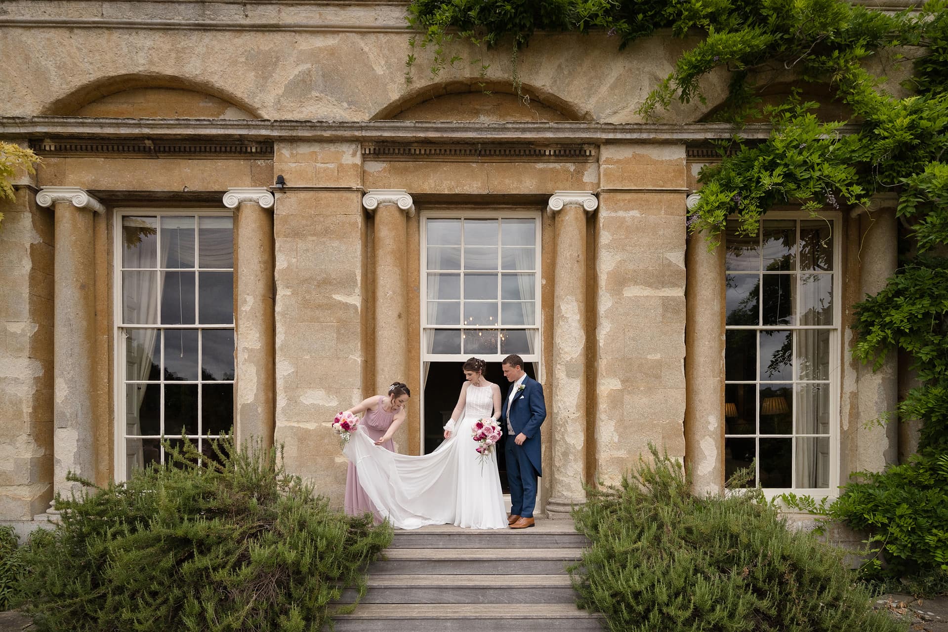 Bridesmaid arranging the bride's dress as she walks out of a sliding sash window door at a period country house