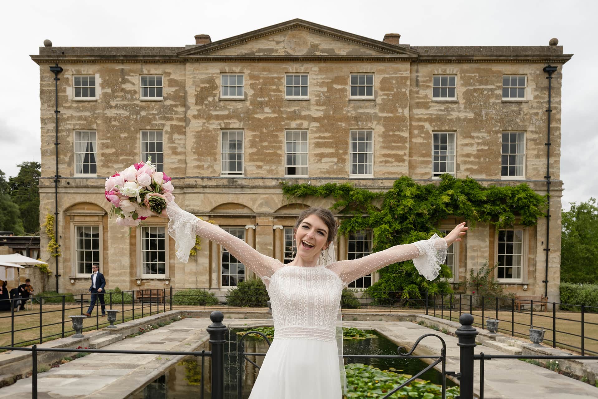 Bride standing with her arms outstretched with Courteenhall House behind her - as if to welcome everyone