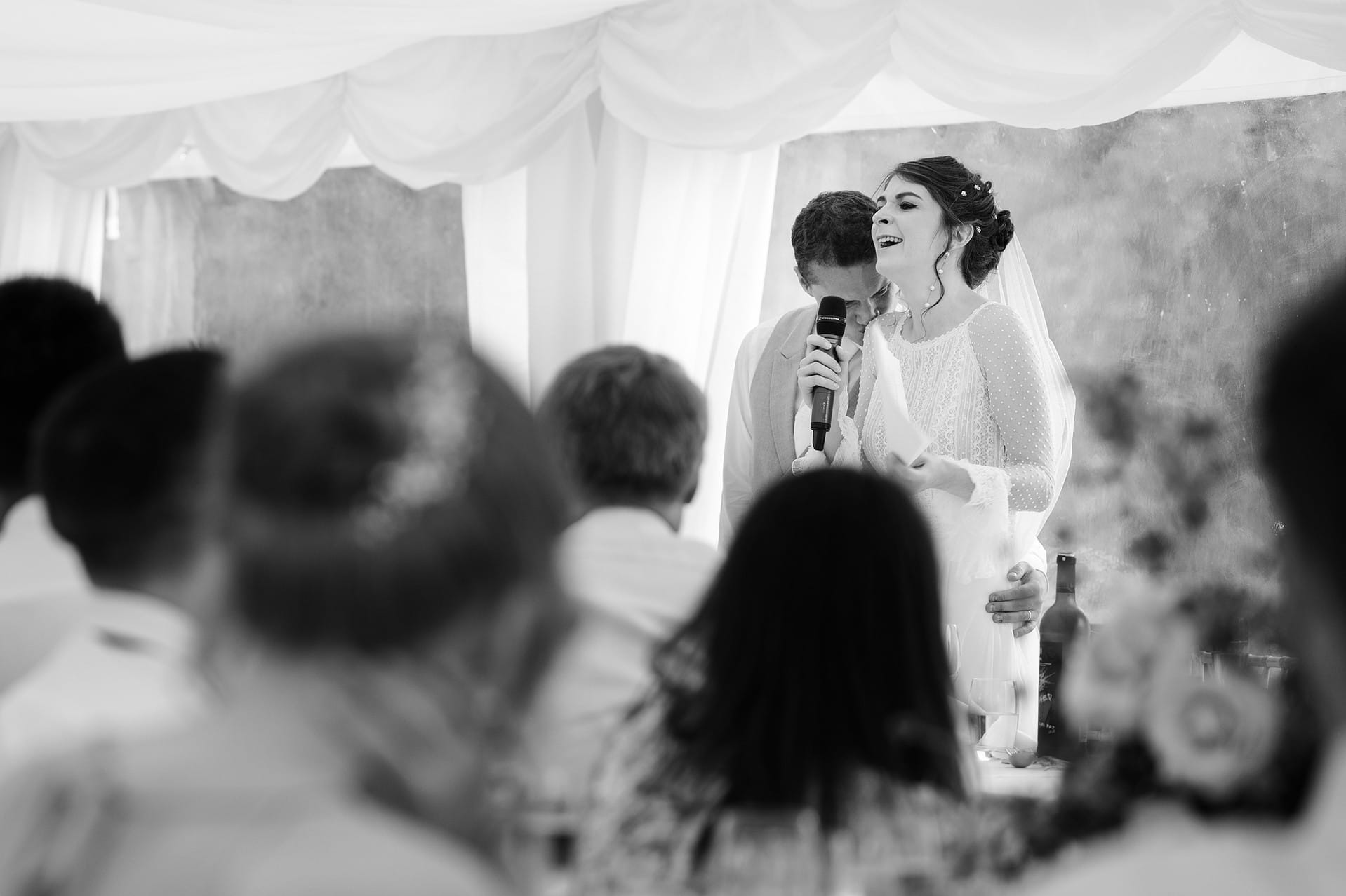 Groom kissing the bride's shoulder as she throws her head back laughing during their joint speech