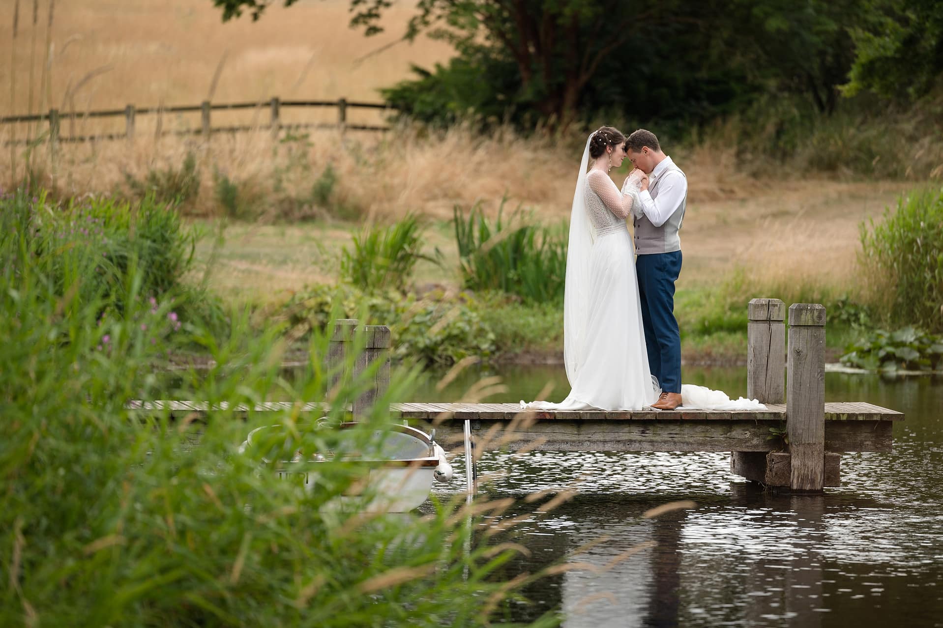 Groom kissing the bride's hands as they stand on a little wooden jetty over a lake at Courteenhall