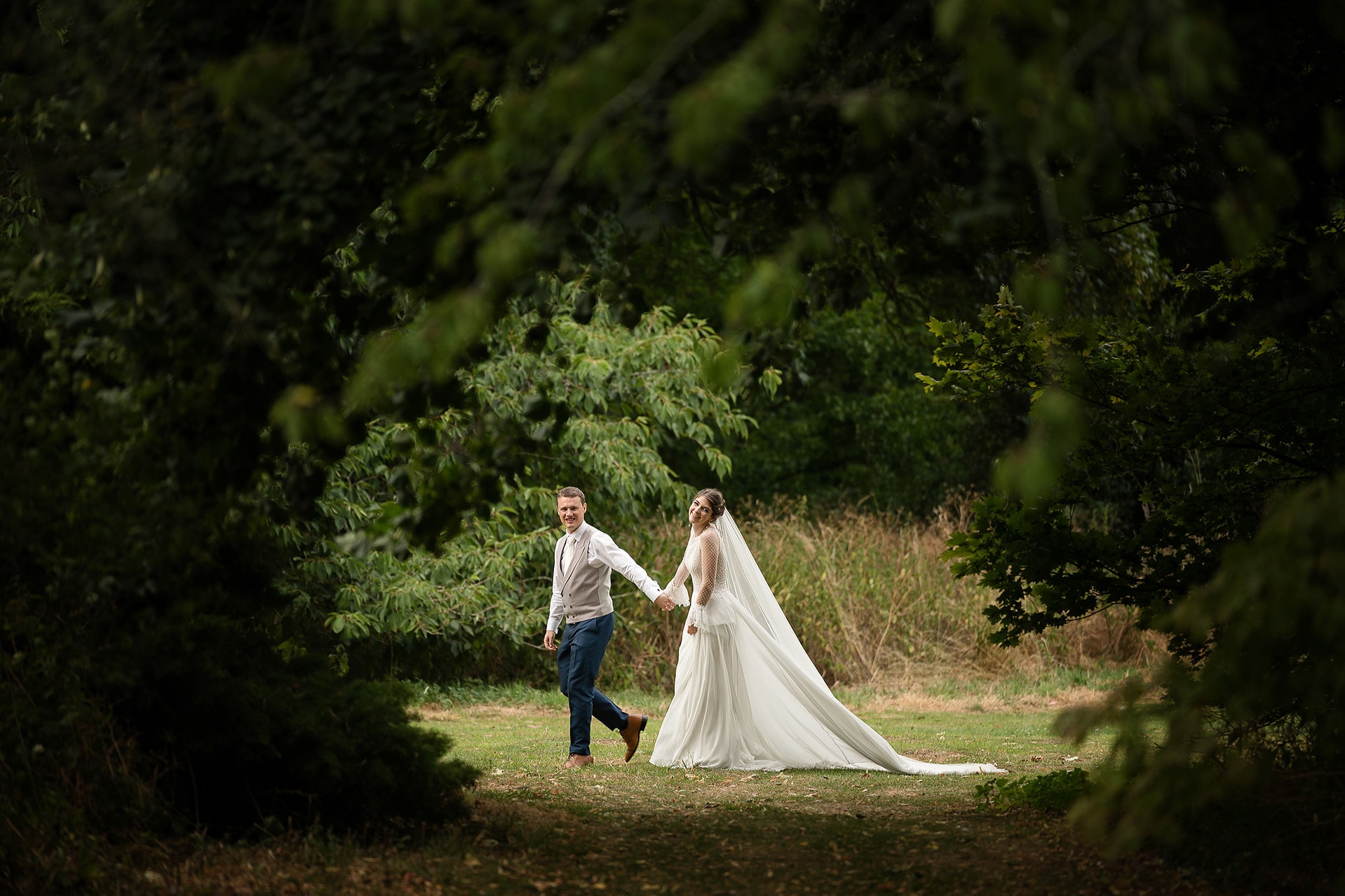 Bride and groom walking hand in hand framed by a gap between the trees in the aboretum at Courteenhall