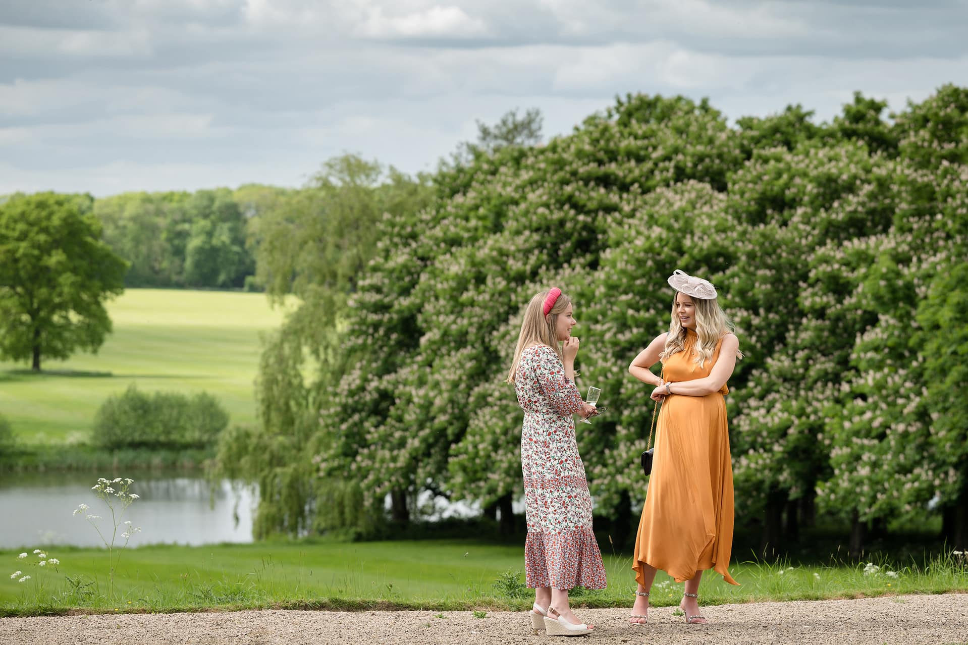 Two wedding guests in colourful dresses and hats chatting during the drinks reception with the Kelmarsh Hall lake and trees behind them